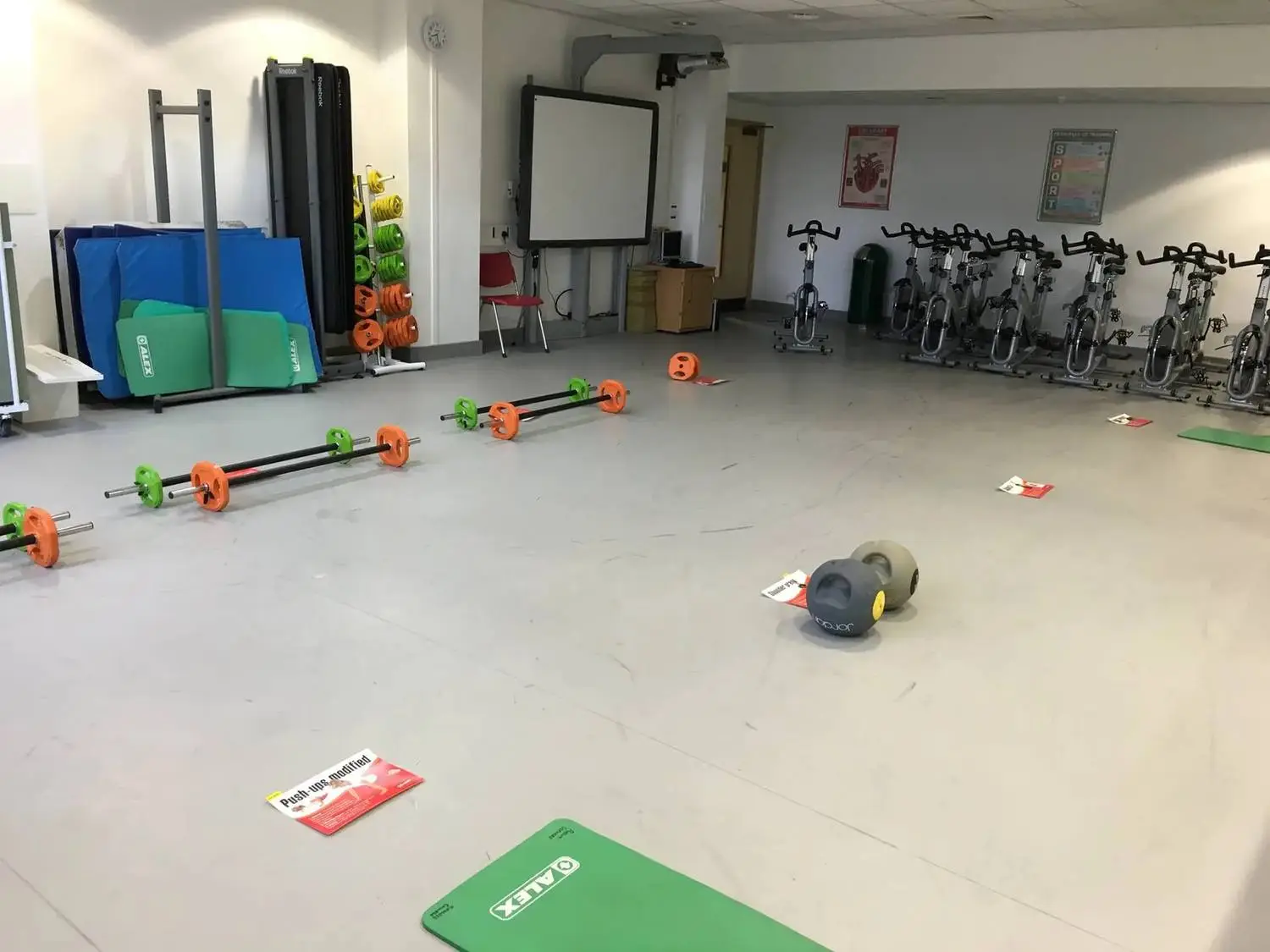 college gym with exercise bikes, barbells, medicine balls, and mats arranged for a fitness class.