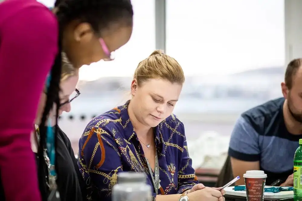 A woman in a blue patterned shirt sits at a table, focused on writing in a notebook, with two others nearby in a classroom setting.