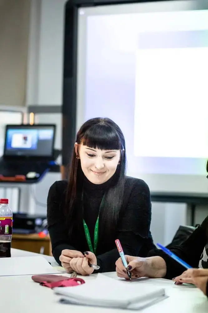 Two students in a classroom, one writing in a notebook, both focused on their work, with a projector screen in the background.