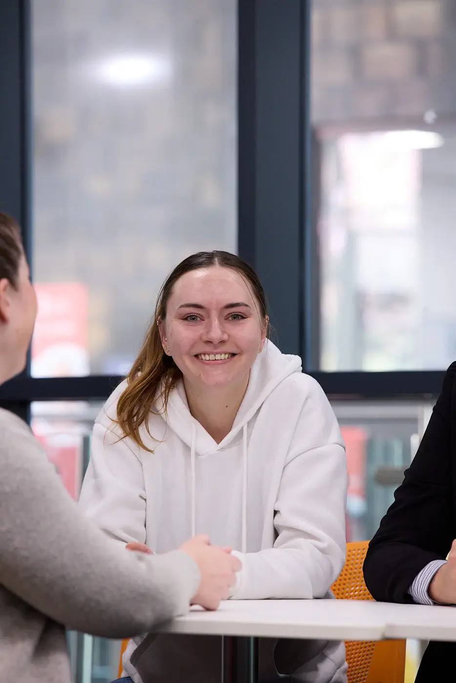 A smiling student in a white hoodie sits at a table in a brightly lit college room, engaged in conversation with two others.