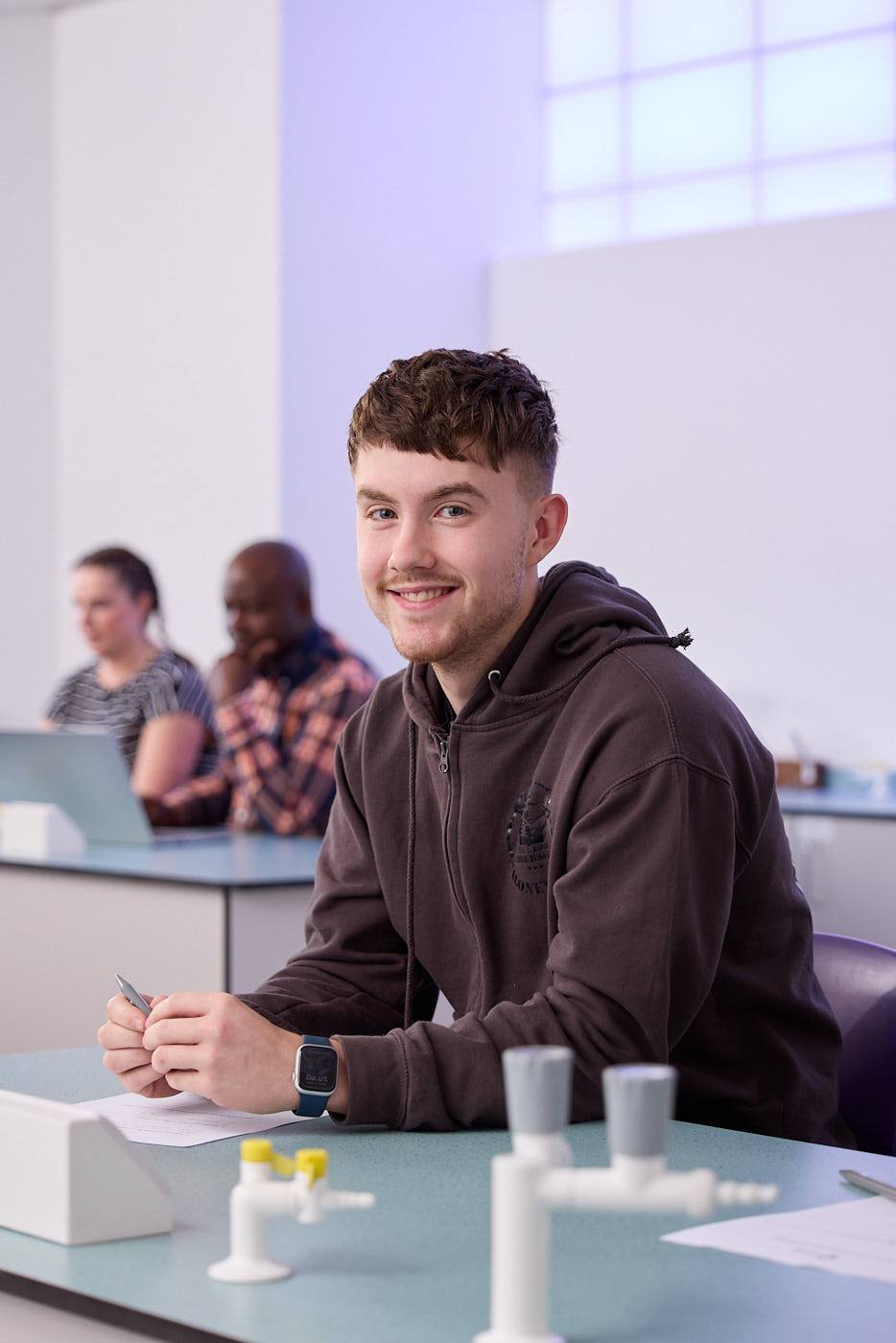 A student in a brown hoodie smiles at the camera while sitting at a lab table with scientific equipment and taking notes.