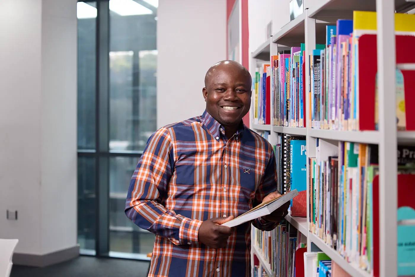 A man in a checkered shirt stands in a library, holding a book and smiling at the camera.