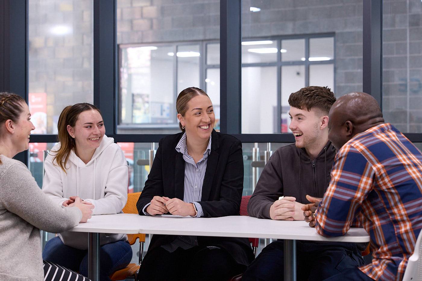 A group of students sitting around a table in a college classroom, engaged in a discussion and smiling.
