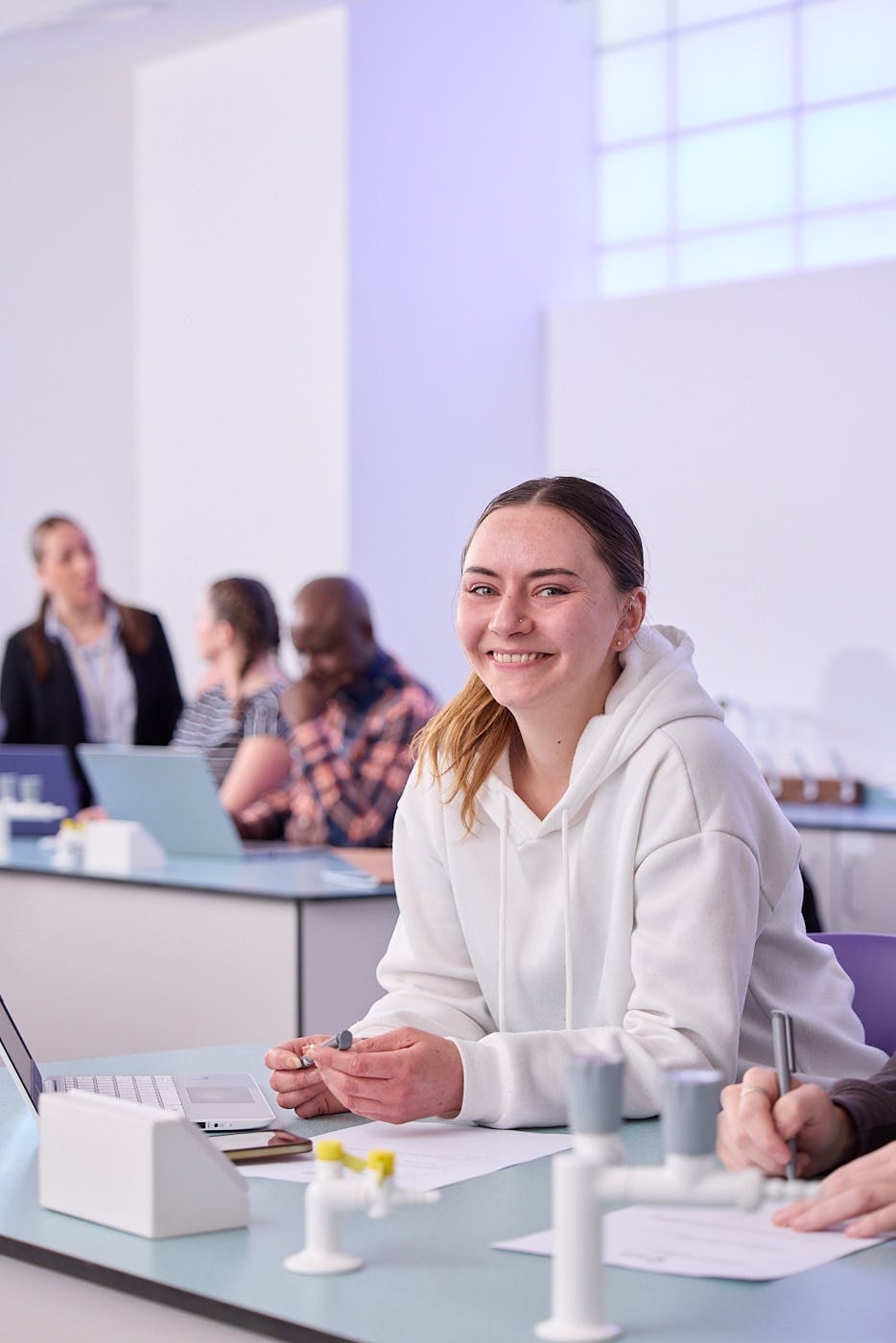 A student in a white hoodie smiles at the camera while sitting at a desk with a laptop and scientific equipment in a brightly lit classroom.