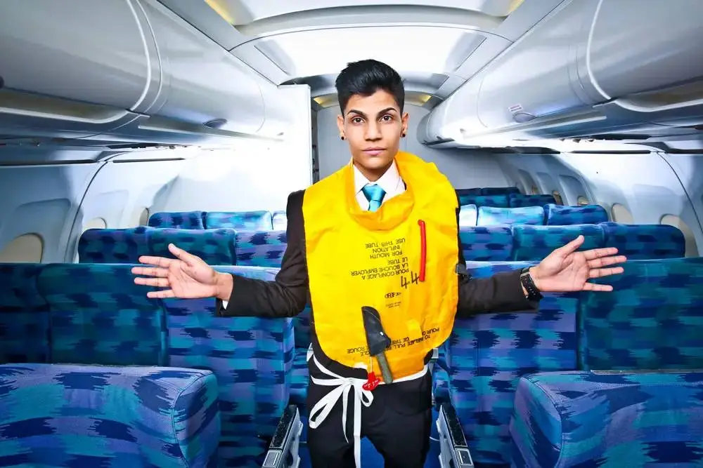 A man in a suit and life jacket stands in an empty aeroplane aisle, demonstrating safety procedures.
