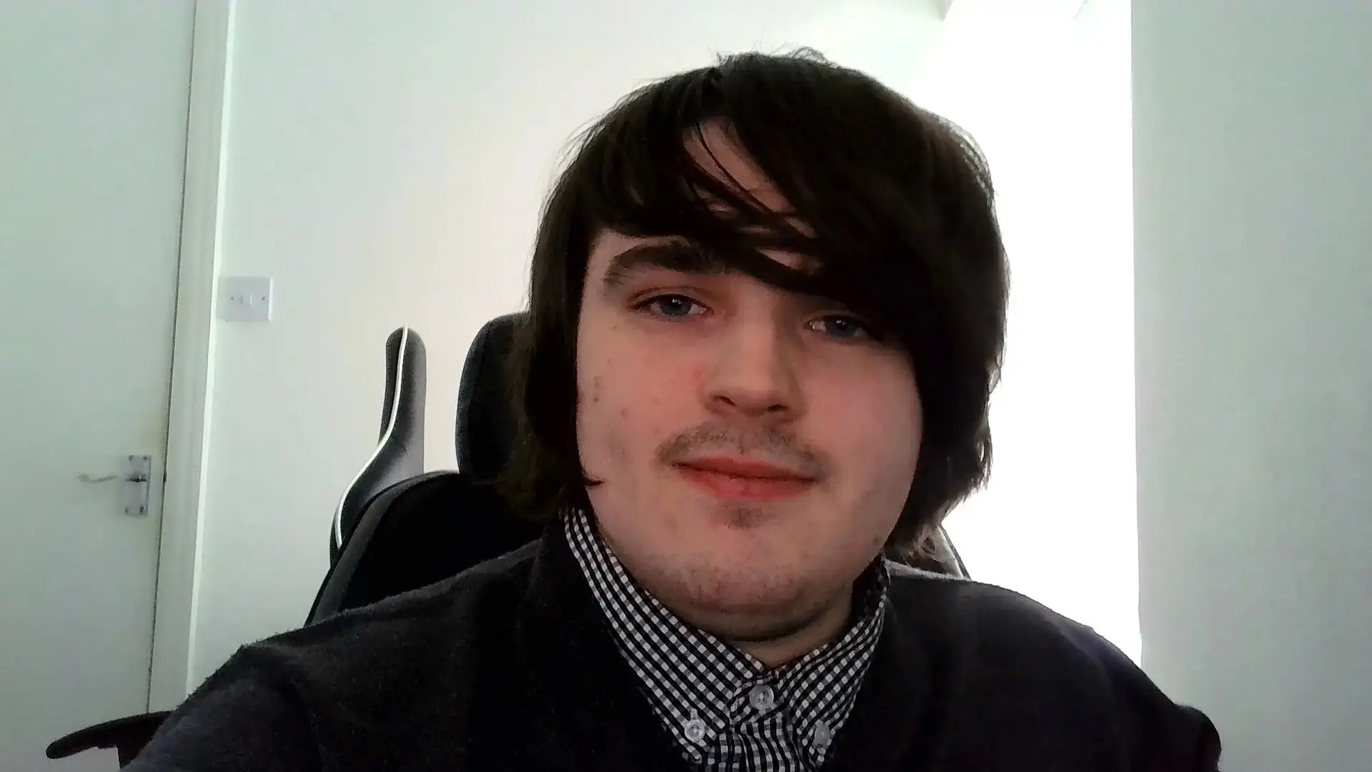 A young man with dark hair and a checkered shirt sits in an office chair, smiling at the camera.