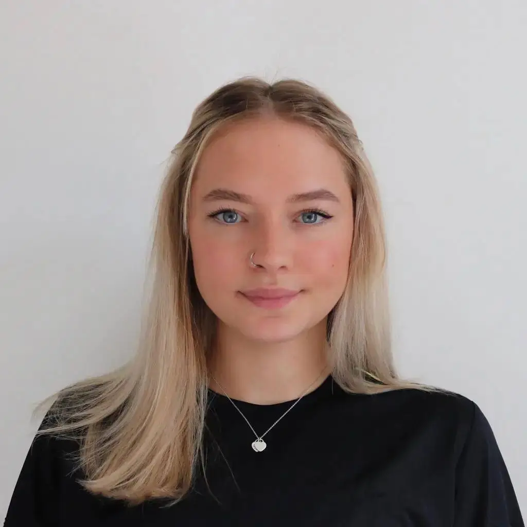 A young woman with long blonde hair and a nose ring poses for a photo against a white background. She wears a black top and a silver necklace with a heart-shaped pendant.
