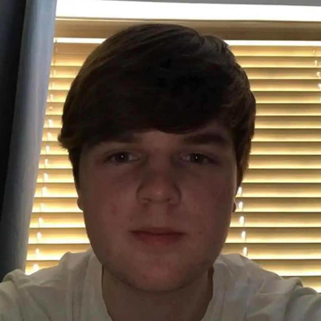 A young man with short dark hair poses for a photo in front of a window with blinds.