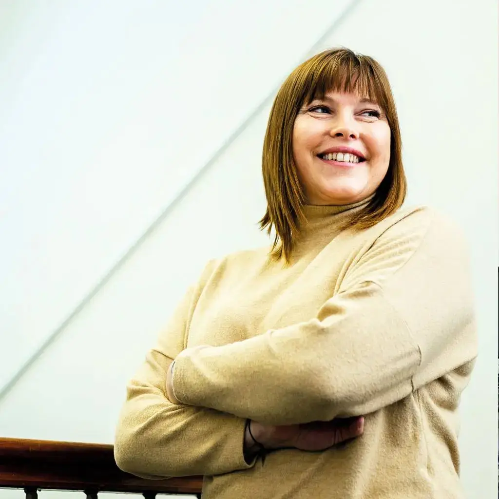 A woman in a beige jumper stands with arms crossed, smiling and looking upwards. She has shoulder-length brown hair with bangs. The background is a white ceiling with diagonal lines.