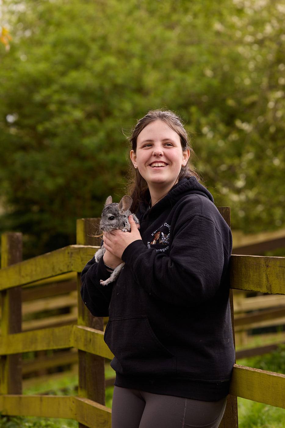 woman in a black hoodie holds a grey rabbit near a wooden fence, smiling in a green outdoor setting.