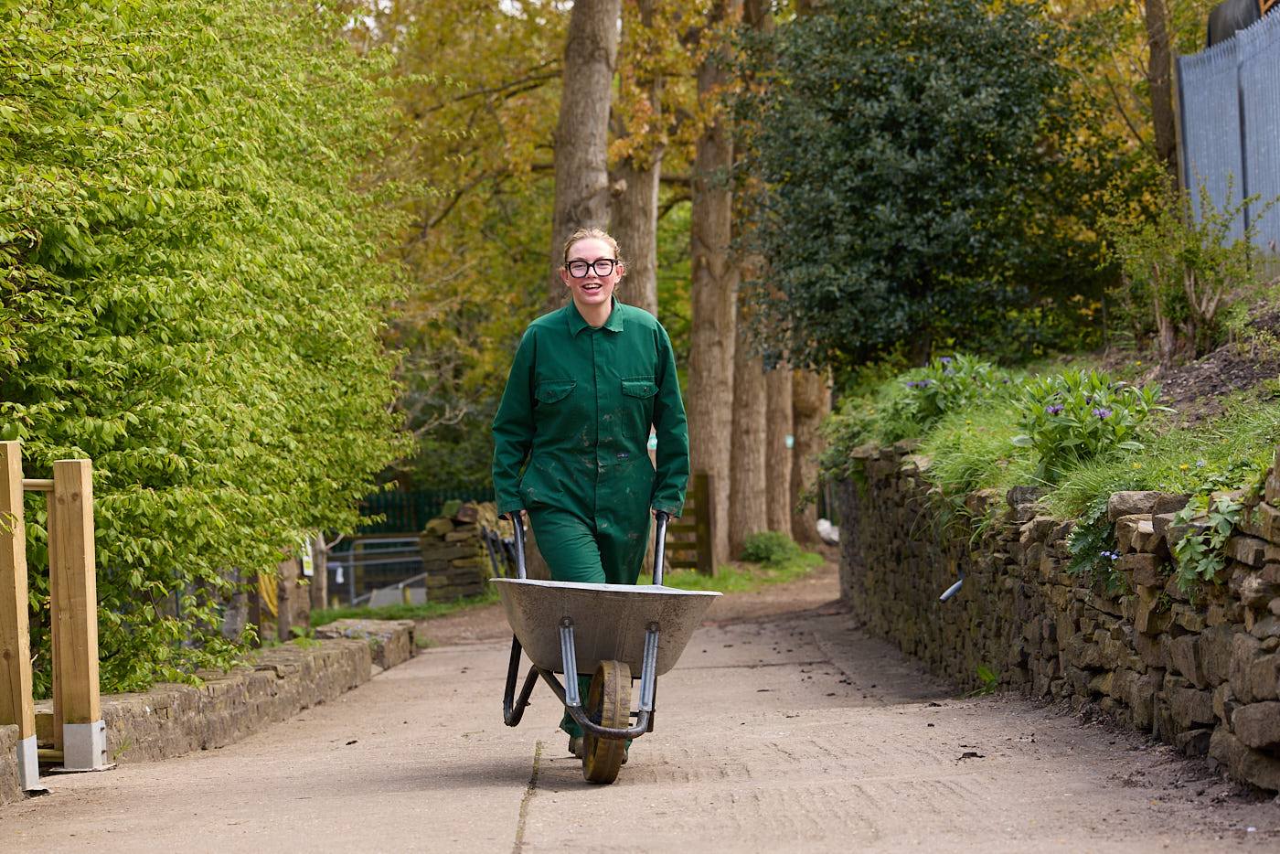 woman in green overalls pushing a wheelbarrow along a tree-lined path on a college campus.