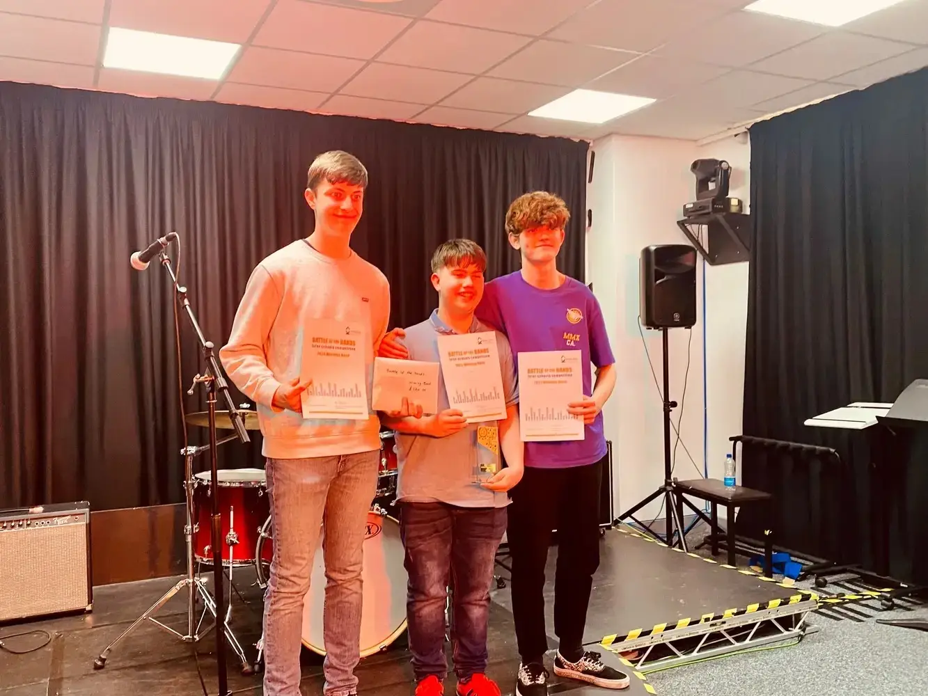 Three students on stage holding certificates, smiling, with musical equipment and a microphone in the background. Educational achievement celebration.