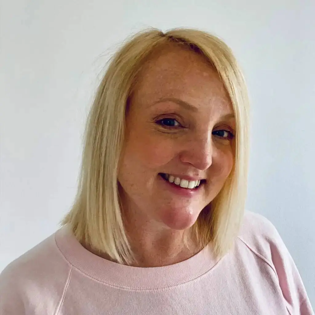A smiling woman with blonde hair in a pink top, standing against a white background. She appears to be in a professional or educational setting.