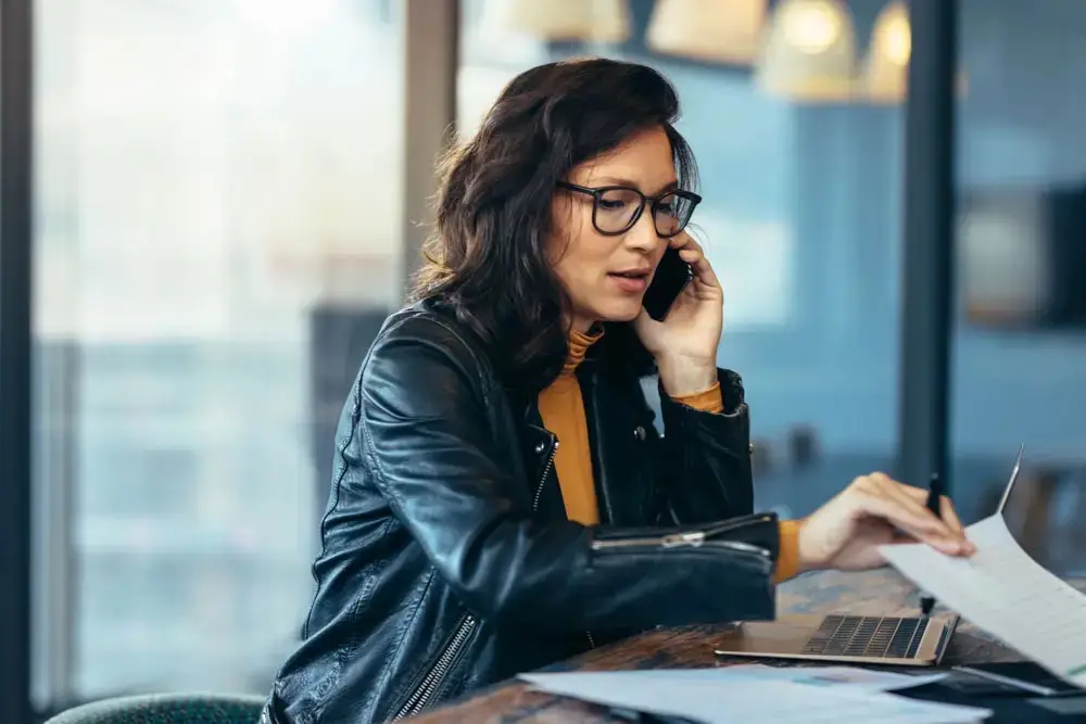A woman in a black leather jacket talks on the phone while working on a laptop and holding papers in a brightly lit room.