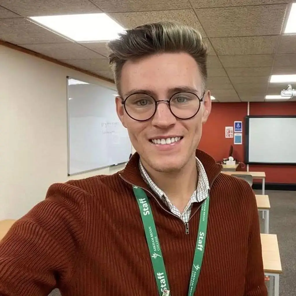 A man in a brown jumper and glasses smiles in a classroom with a whiteboard and projector screen.