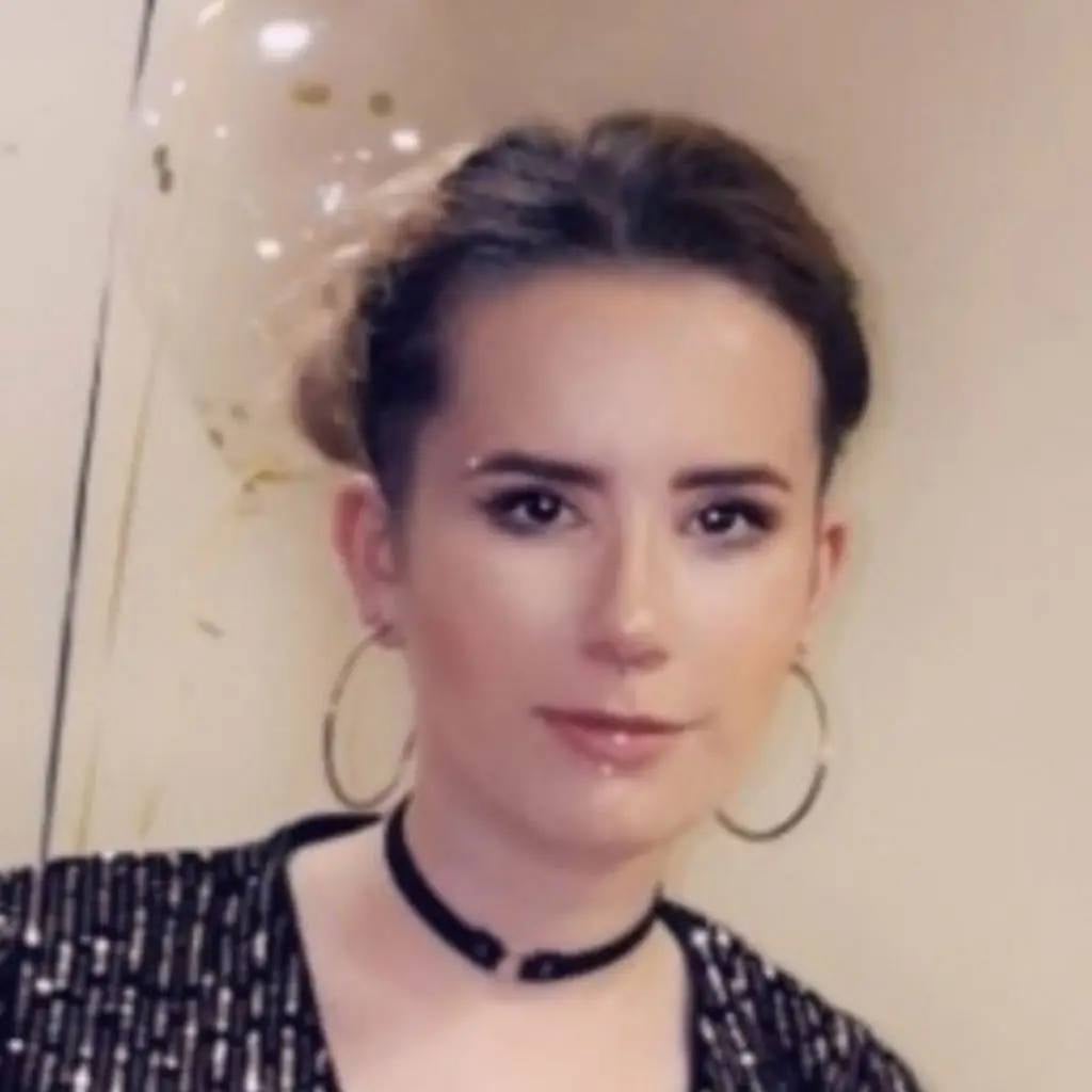 woman with dark hair styled in a bun wears a black choker and hoop earrings. She stands against a light background, exuding a confident and poised appearance.