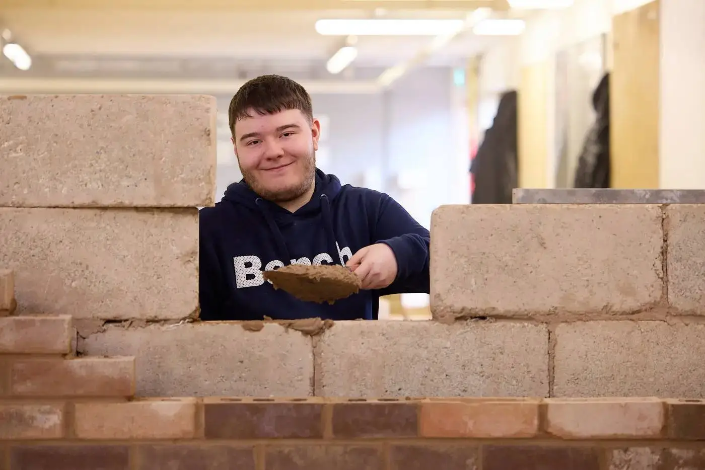 A student in a blue hoodie smiling and holding a trowel while building a wall with concrete blocks in a classroom.