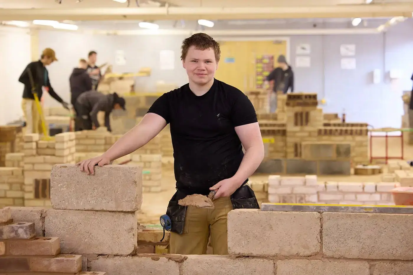 A student in a black t-shirt building a brick wall in a workshop with other students in the background.