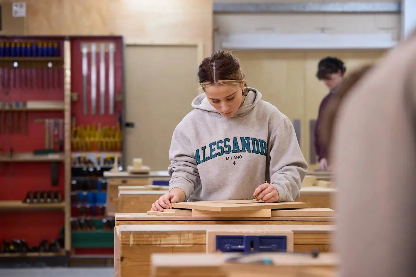 A student in a grey hoodie working on a wooden project in a well-equipped college workshop.