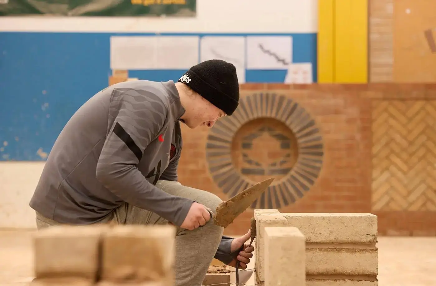 A student in a beanie kneels, using a trowel to spread mortar on a concrete block in a workshop setting.