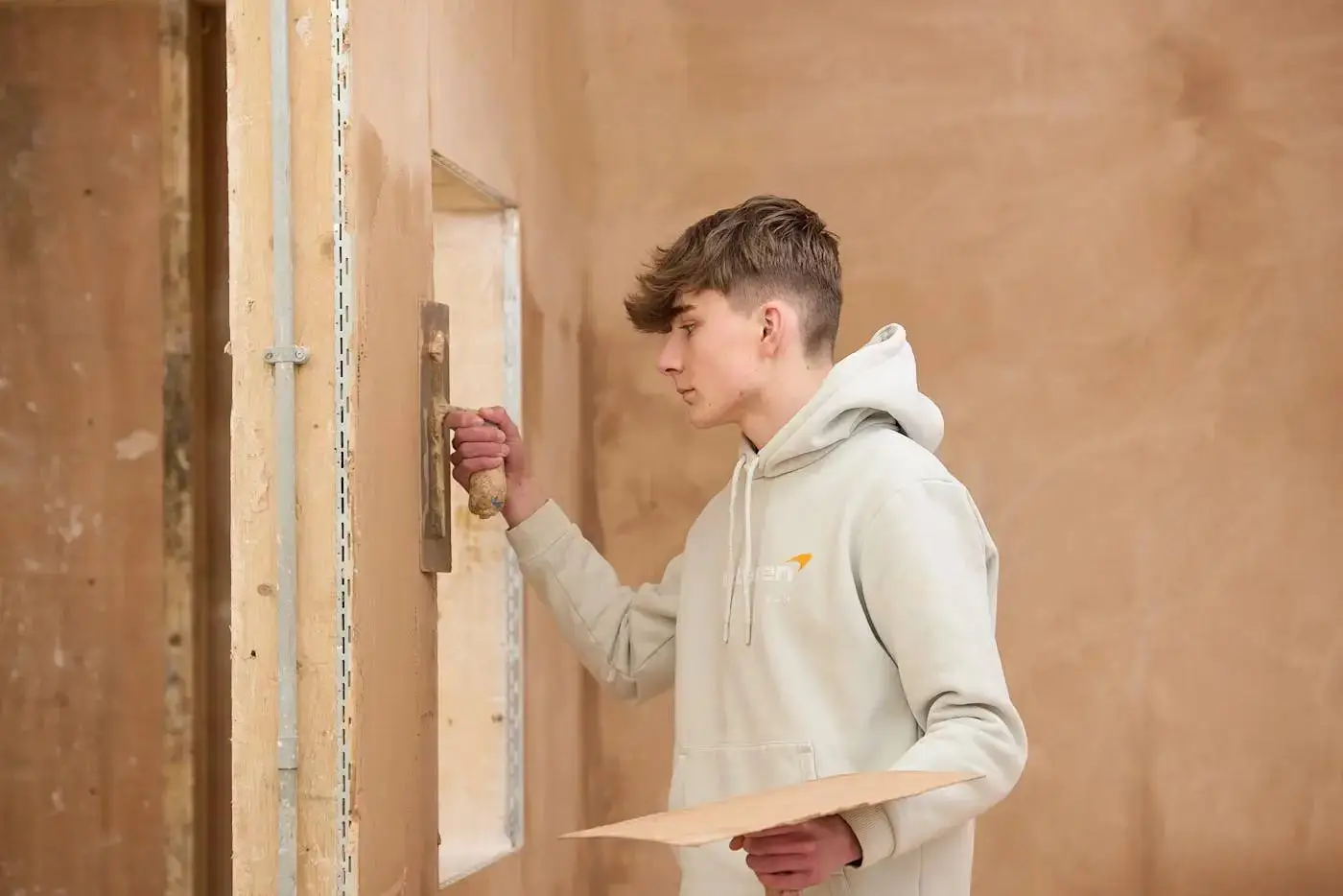 A student in a white hoodie applies plaster to a wall with a trowel, demonstrating practical construction skills in a workshop setting.