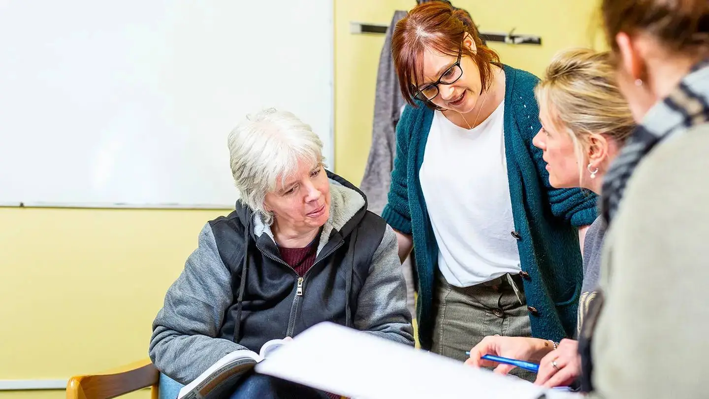 A group of people in a classroom setting, engaged in discussion around an open book. One person is seated, while others stand nearby, smiling and attentive.