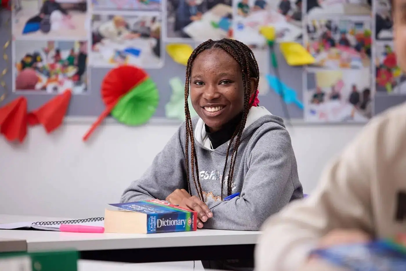 A student smiling in a classroom, sitting at a desk with a dictionary and notebook.