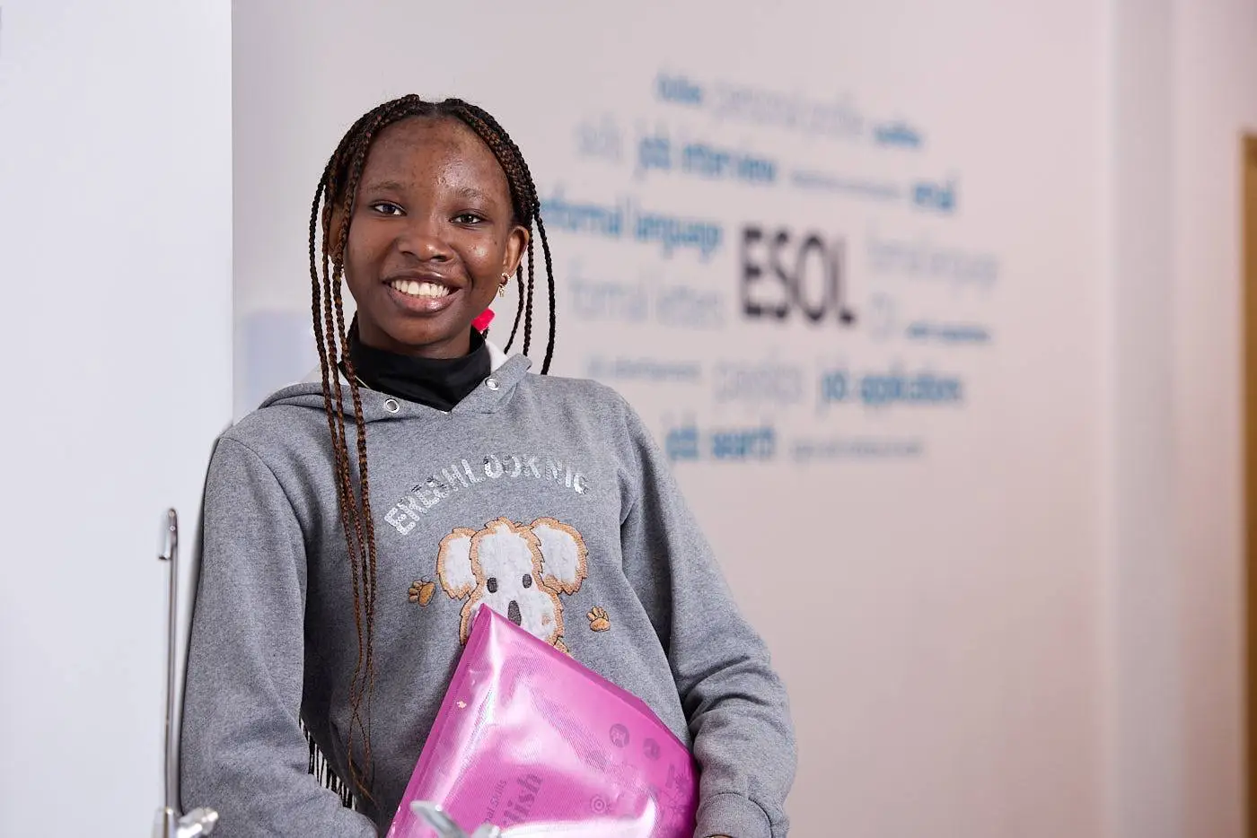 A smiling student in a grey hoodie holding a pink folder stands in front of a wall with English language words.