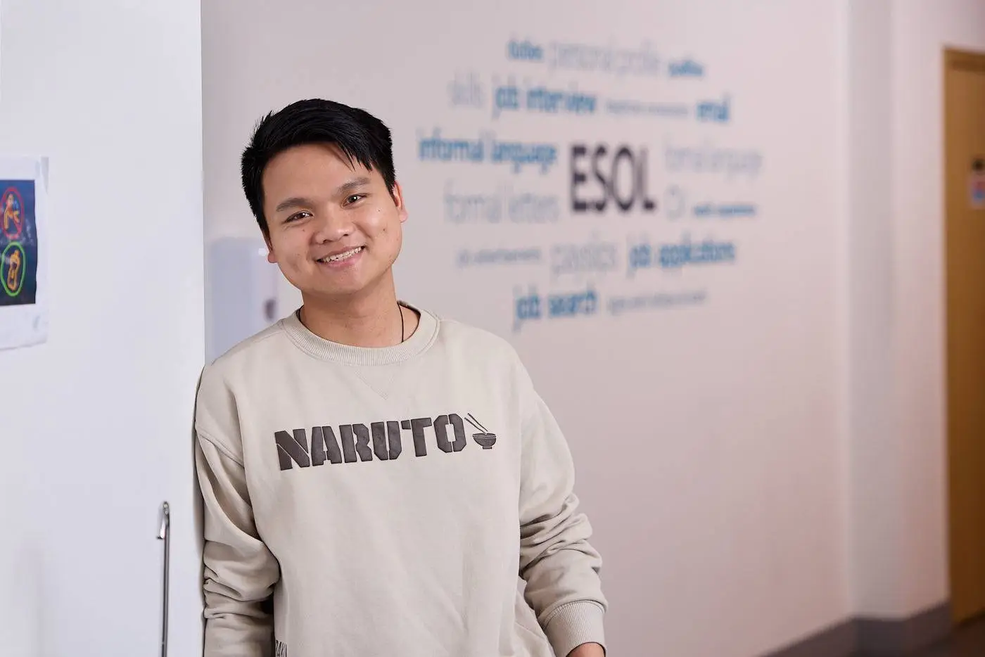 A young man in a beige sweatshirt smiling in front of a wall with English language learning words.