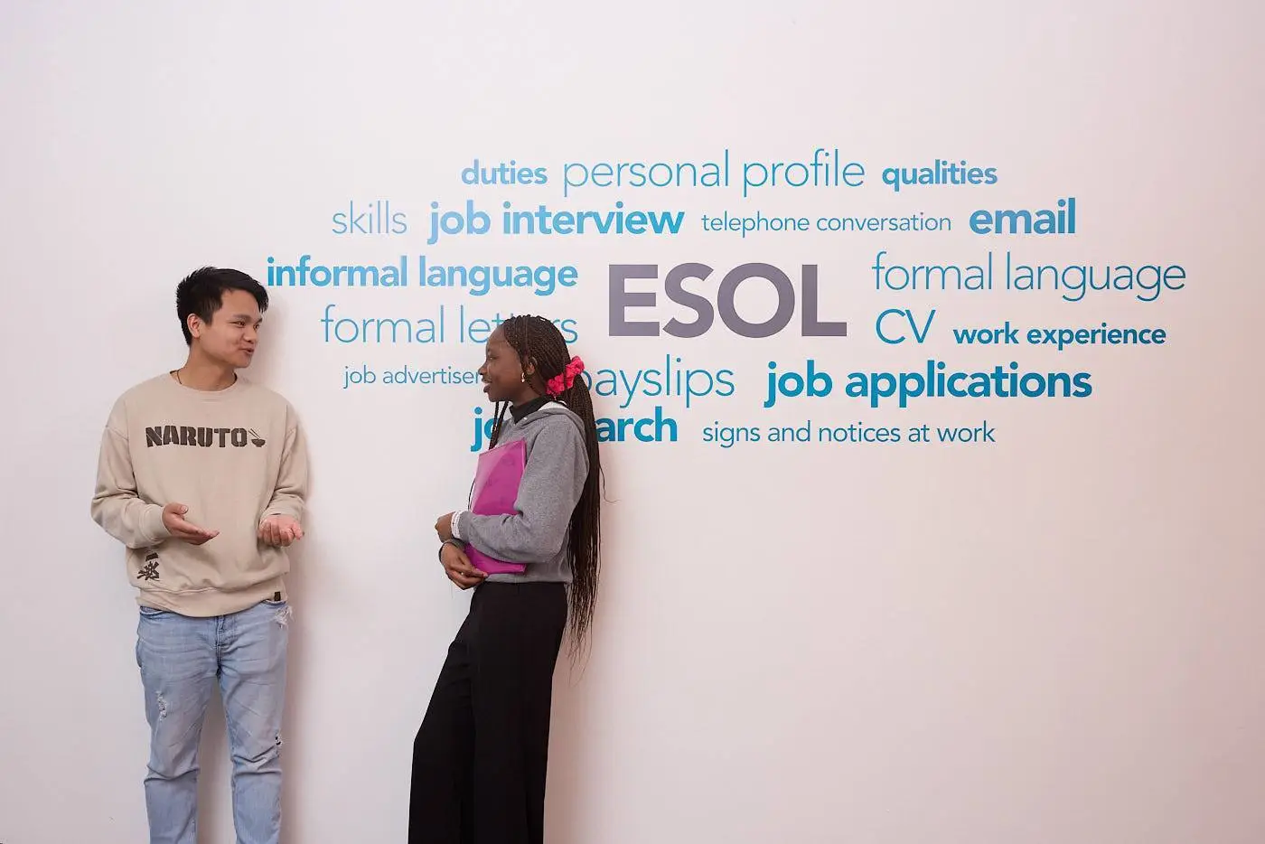 Two students conversing in front of a wall with job-related terms like 'ESOL', 'CV', 'interview', and 'formal language', highlighting employment skills development in a college setting.
