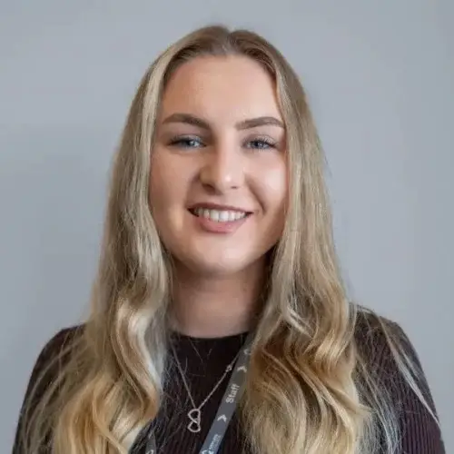 Smiling woman with long blonde hair wearing a lanyard and necklace, standing against a grey background. Suitable for a college profile or staff introduction.