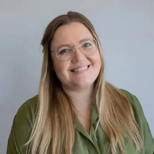 A smiling woman with long blonde hair and glasses wearing a green shirt, standing against a grey background.