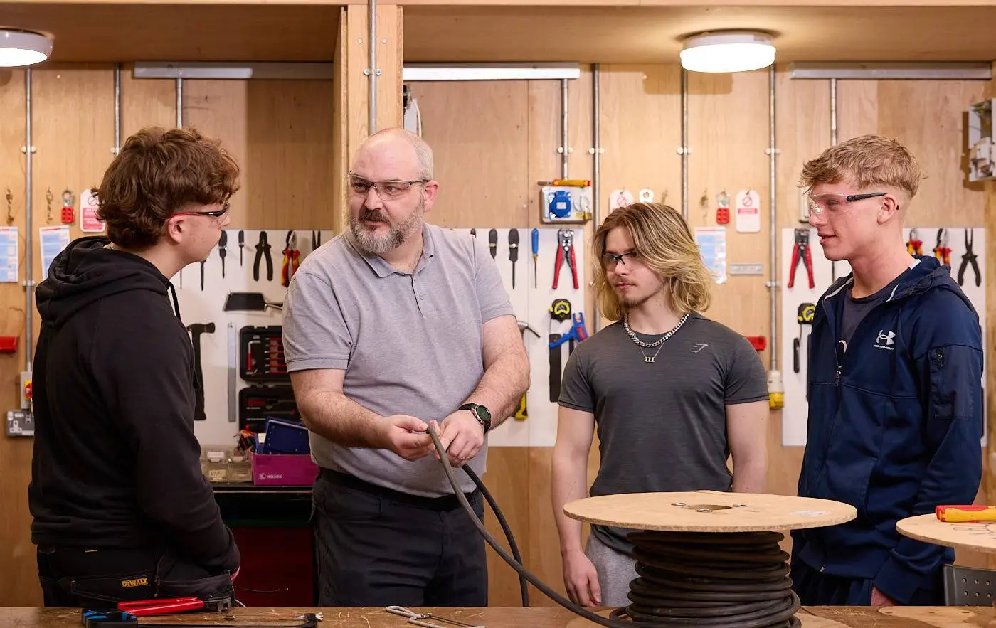 A man in a grey polo shirt teaching three students in a workshop with tools and equipment on the walls.