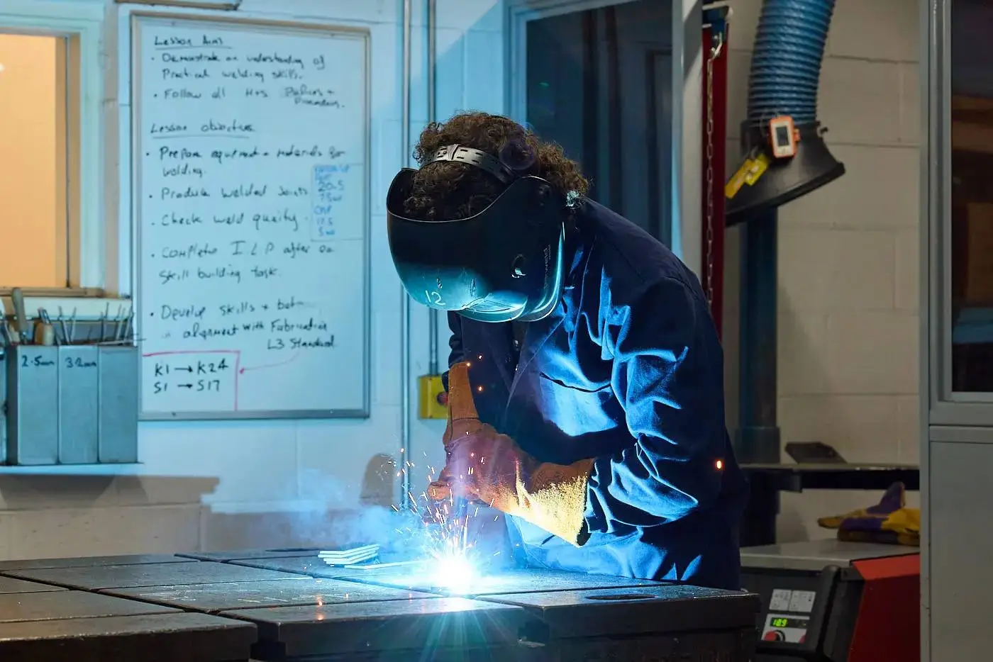 A student in protective gear welding metal in a workshop, with a lesson plan on the wall.