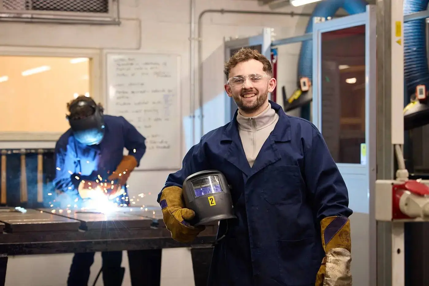 A student in a welding lab, wearing protective gear, smiling and holding a welding helmet, with sparks flying from a welding torch in the background.