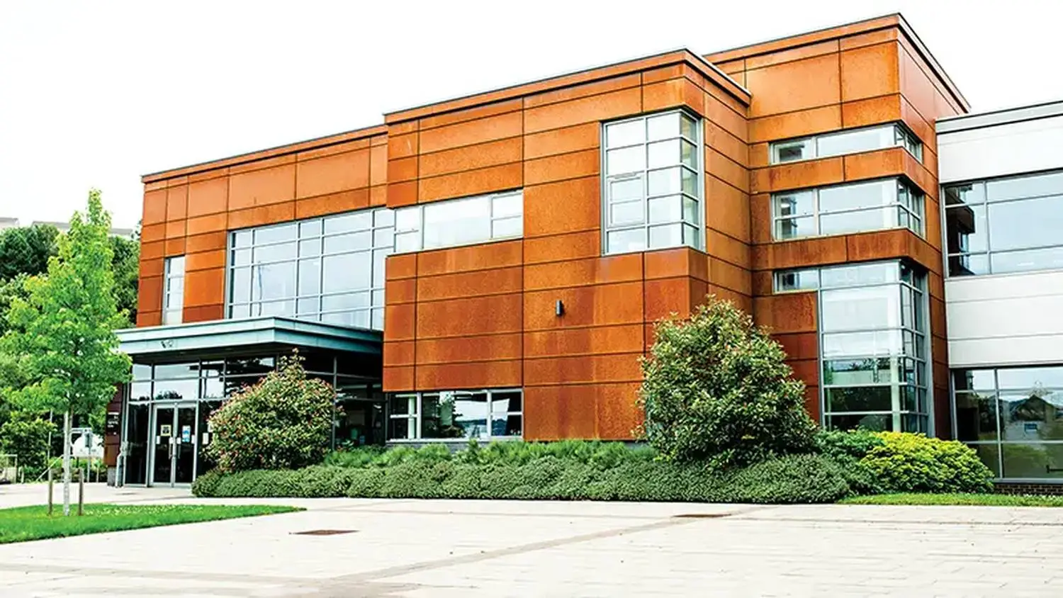 Modern college building with orange and glass facade, entrance with sliding doors, and landscaped pathway under a partly cloudy sky.