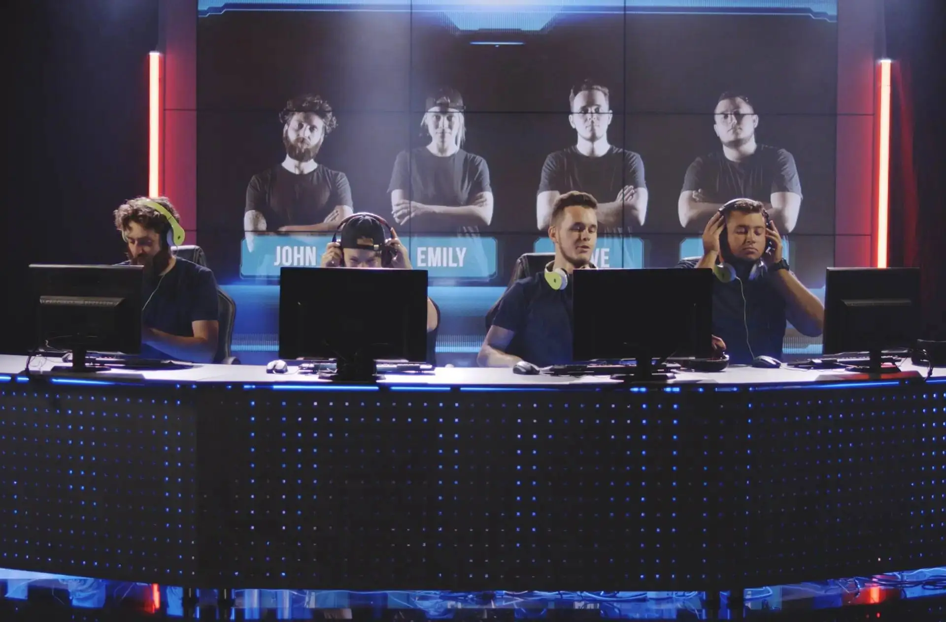 Four students in headphones at computer desks on a stage with a large screen behind them displaying names.