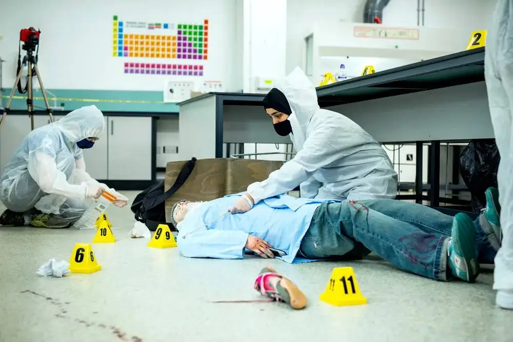 Two forensic scientists in white suits examine a mannequin at a crime scene simulation in a laboratory.