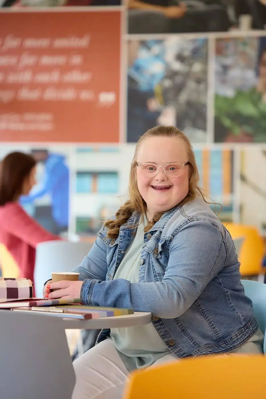 A smiling student in a denim jacket sits at a table with books and a coffee cup in a brightly coloured college cafe.
