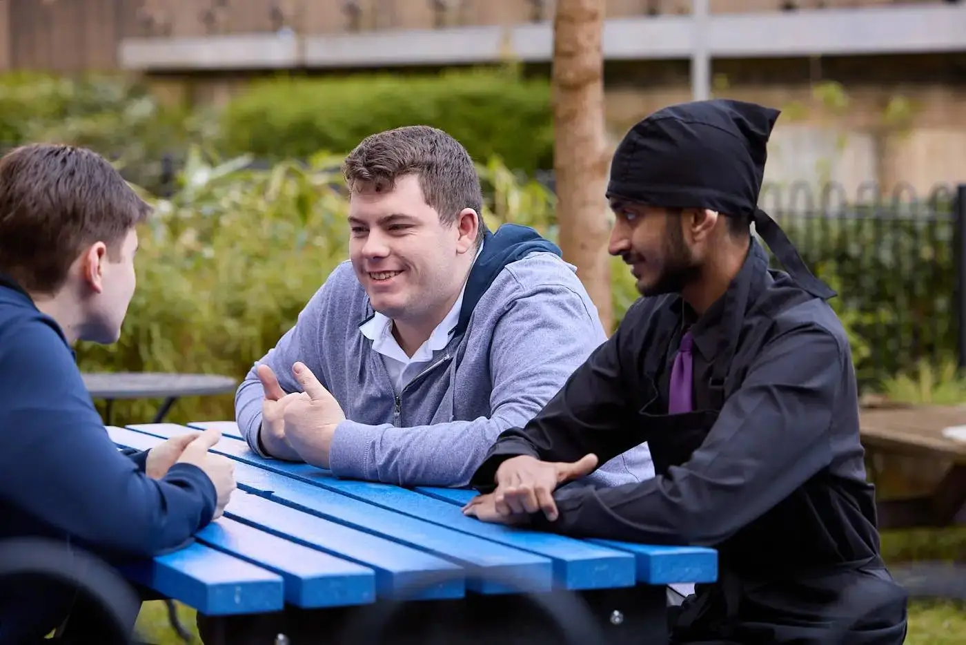Three students sitting at a blue picnic table outdoors, engaged in conversation, with one gesturing animatedly.