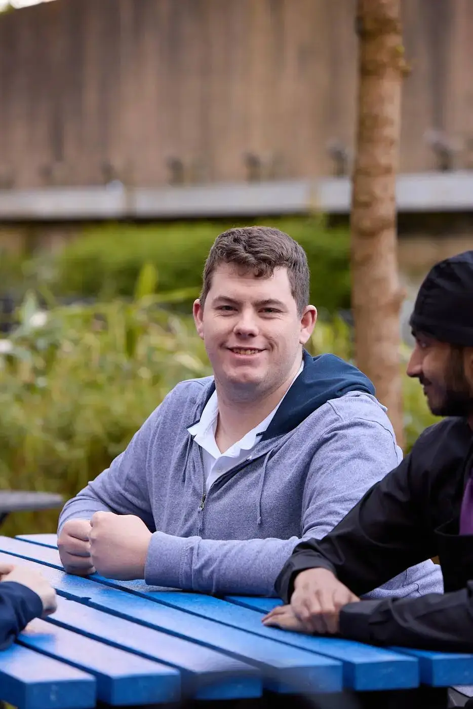 Two men sitting at a blue picnic table outdoors, engaged in conversation. One man is smiling broadly.
