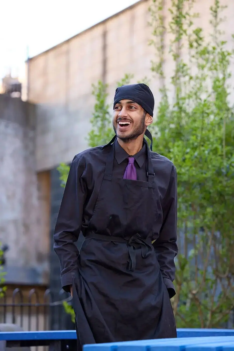 A man in a black chef's uniform and headscarf stands outdoors, smiling and looking to the side. He wears a black shirt and a purple tie. Green plants and a brick wall are in the background.