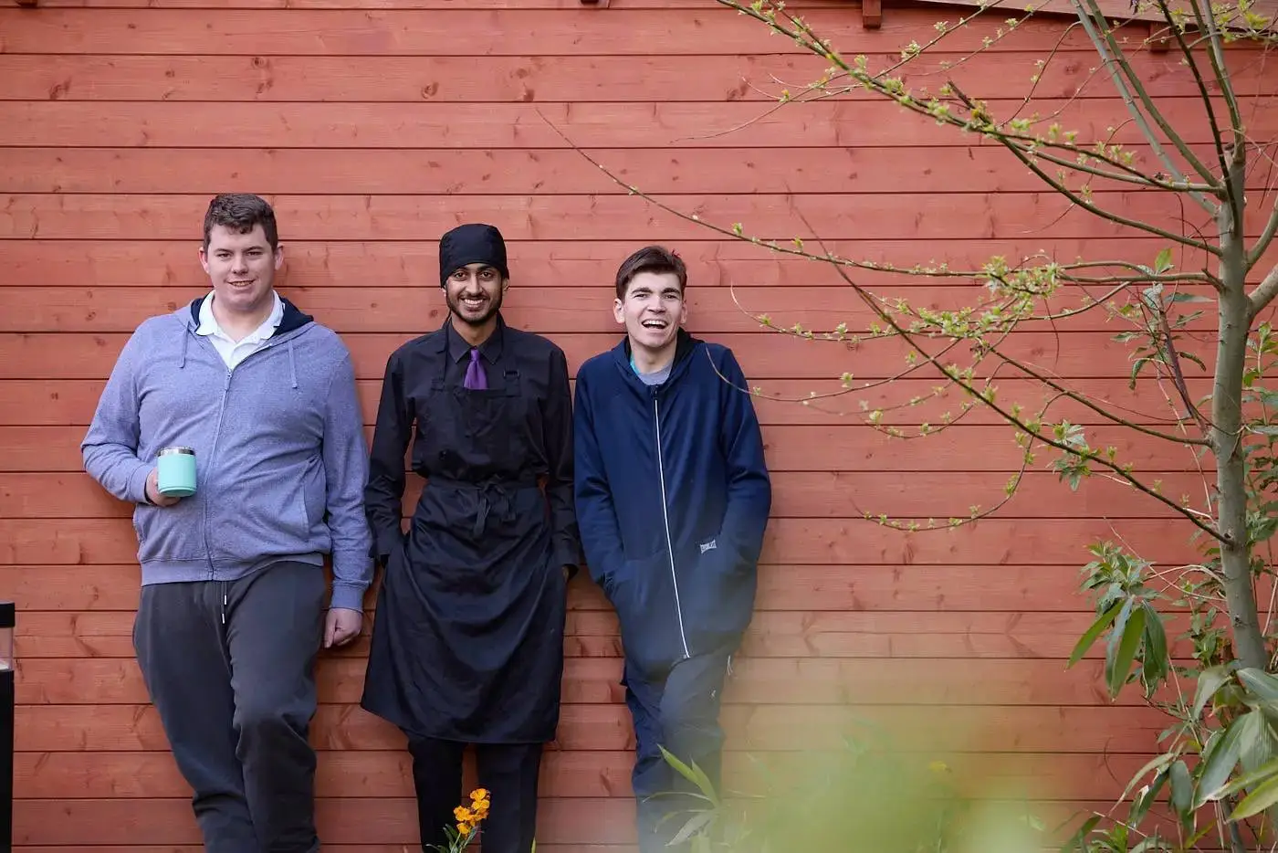 Three men in casual and chef attire stand in front of a red wooden wall, smiling and posing for a photo.