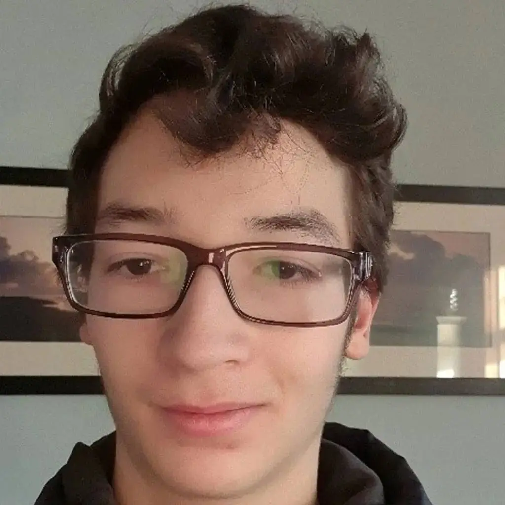 A young man with glasses and dark hair poses for a photo in front of a framed picture of a lighthouse.