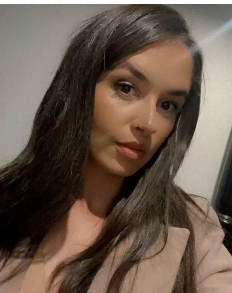 A woman with long dark hair poses for a photo in a well-lit room. She wears a light pink top and looks directly at the camera.