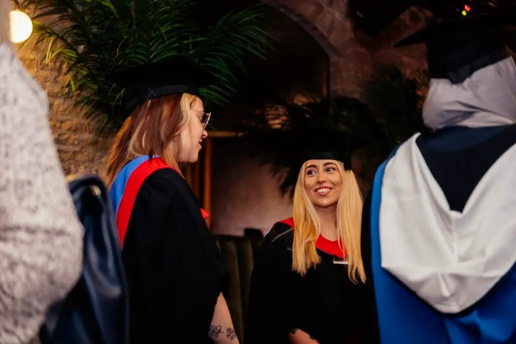 Graduates in black gowns and caps with red and blue hoods, smiling and chatting outdoors at night.