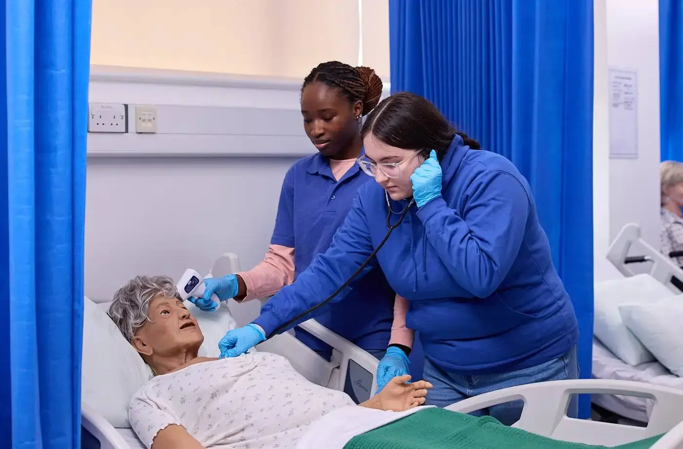 Two healthcare students in blue scrubs and gloves practice using a stethoscope on a hospital mannequin in a clinical setting.