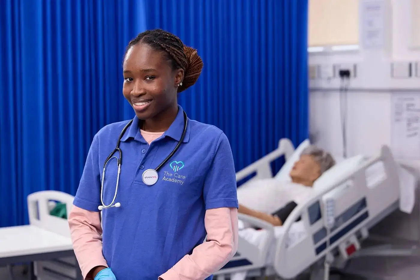 A healthcare student in a blue uniform with a stethoscope stands smiling in a hospital room with a patient in the background.