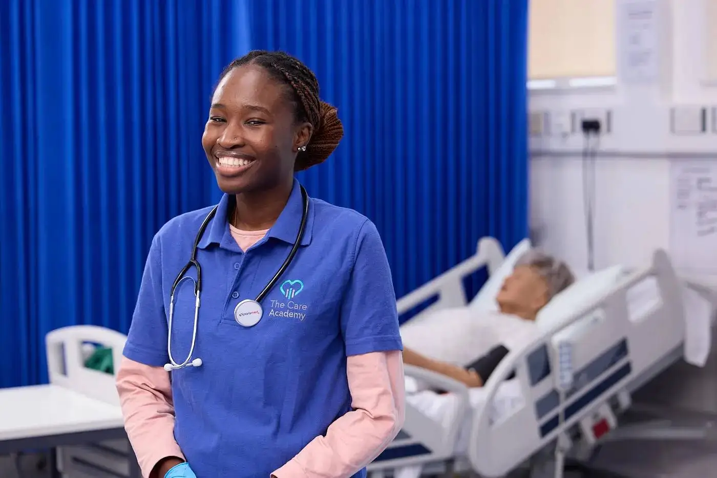 A healthcare student in a blue uniform with a stethoscope stands smiling in a hospital room, next to a patient bed.