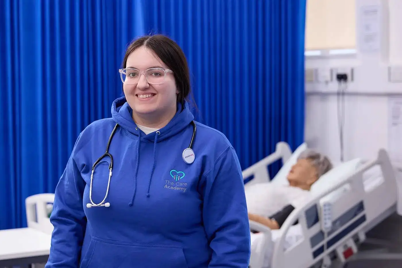 A healthcare student in a blue hoodie with a stethoscope stands smiling in a hospital room, next to a patient bed.